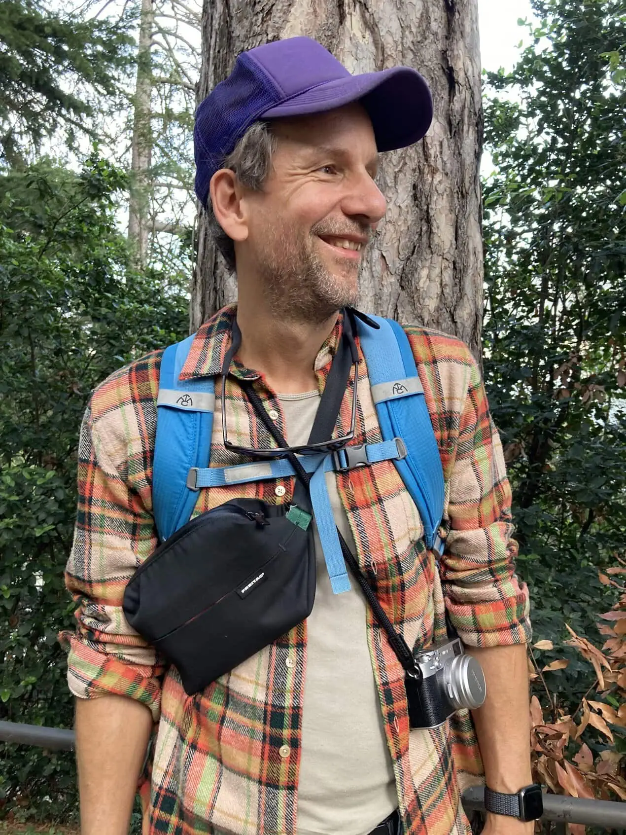 Marcus Smolarek outdoors under a pine tree, wearing a cap, a camera round his neck, smiling off to one side.